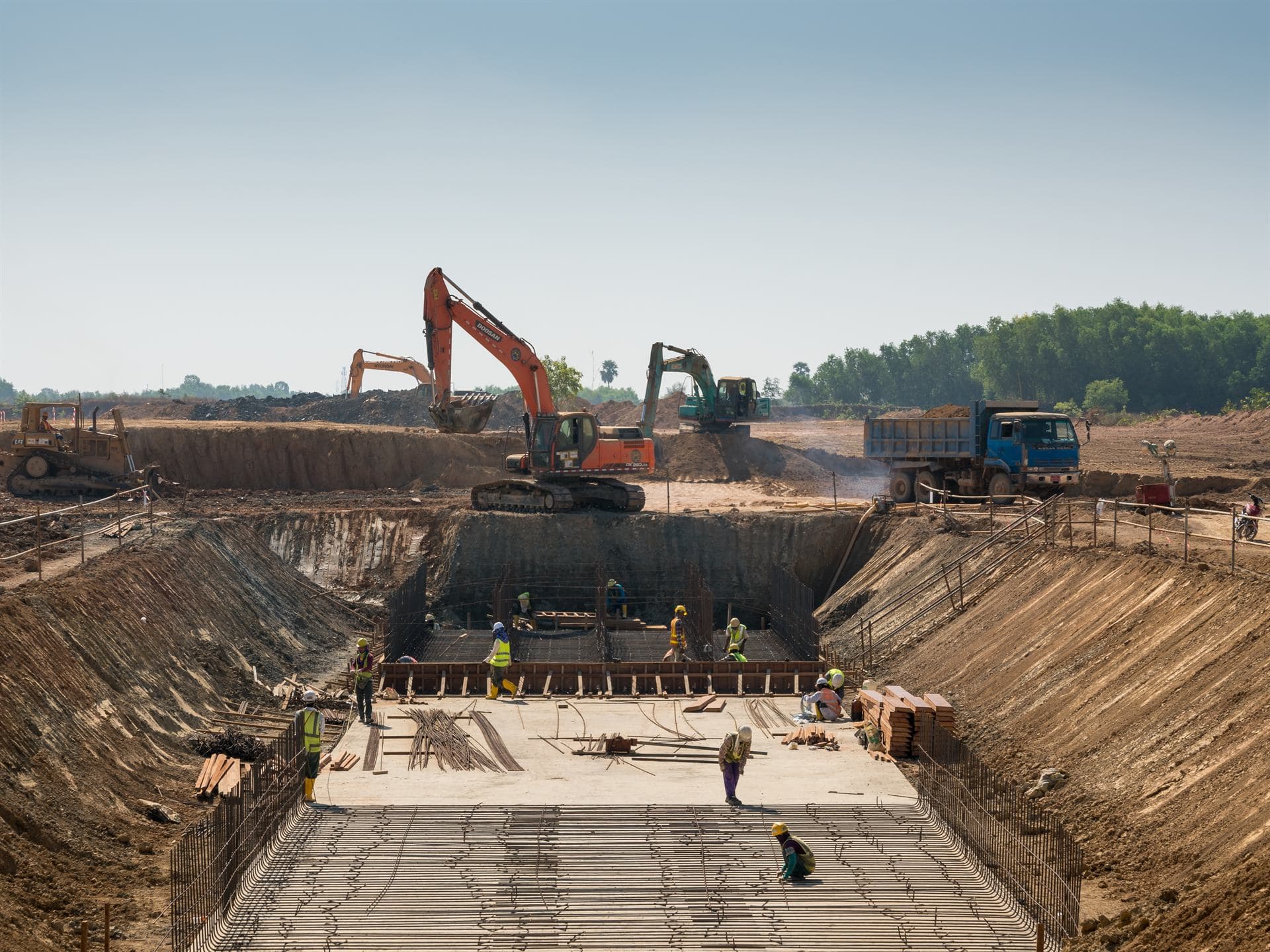Excavación de obra con maquinaria pesada, camión volqueta y obreros armando refuerzo